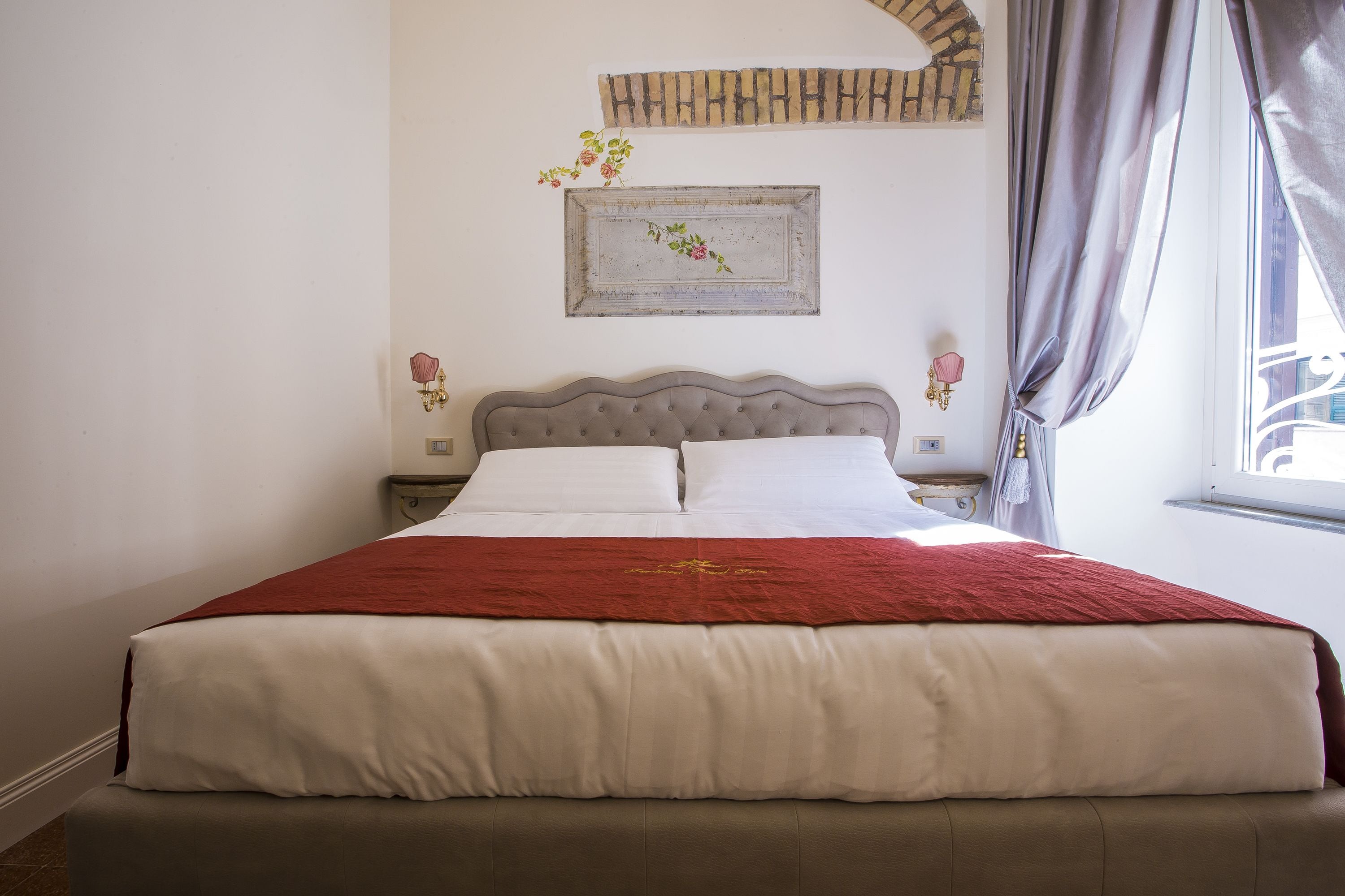 A neatly made bed with a red blanket and white linens, a decorative wall piece above it, and a window with curtains to the right.