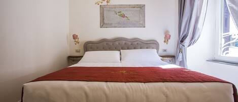 A neatly made bed with a red blanket and white linens, a decorative wall piece above it, and a window with curtains to the right.
