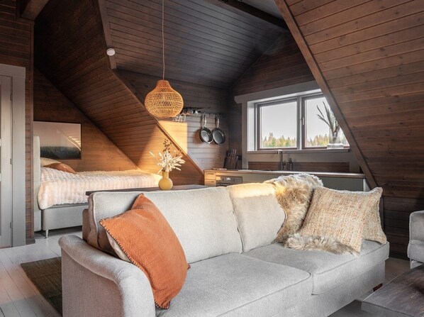 A cozy loft bedroom with a sloped ceiling, a sofa, a bed, and a window overlooking greenery.