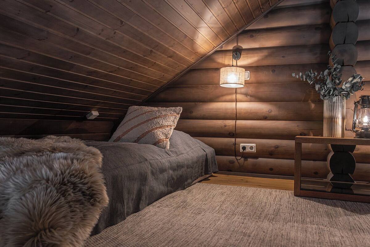Ein gemütliches Schlafzimmer in einer Blockhütte mit schrägem Dach, einem Bett mit flauschiger Decke, einem Nachttisch mit Laterne und einer Wandleuchte.