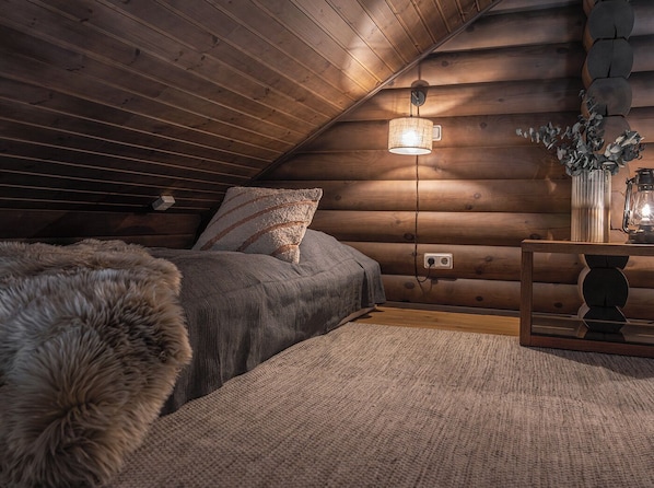 A cozy log cabin bedroom with a sloped ceiling, a bed with a fluffy blanket, a bedside table with a lantern, and a wall-mounted light fixture.