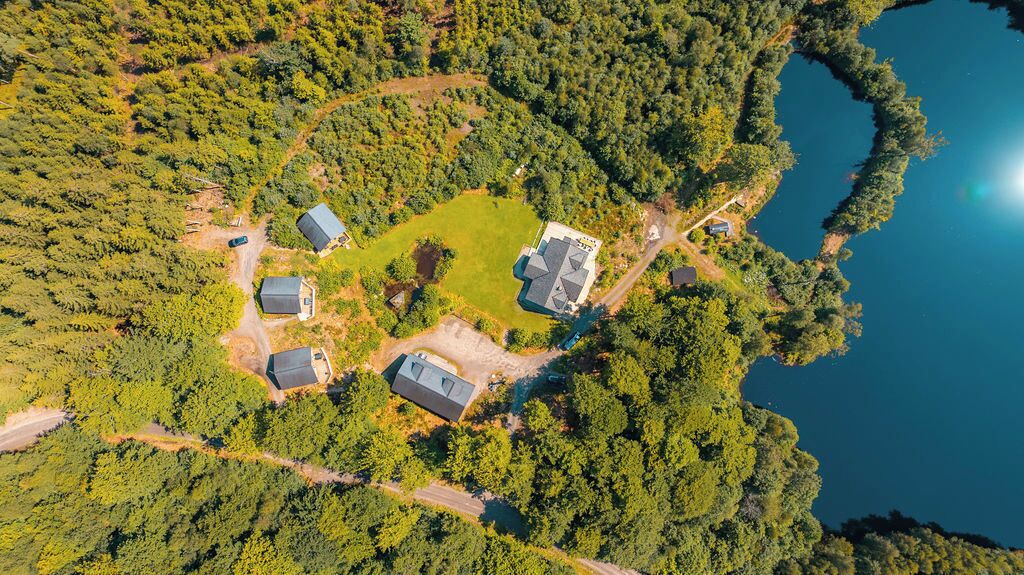 An aerial view of a property with multiple buildings surrounded by dense forest and a lake.