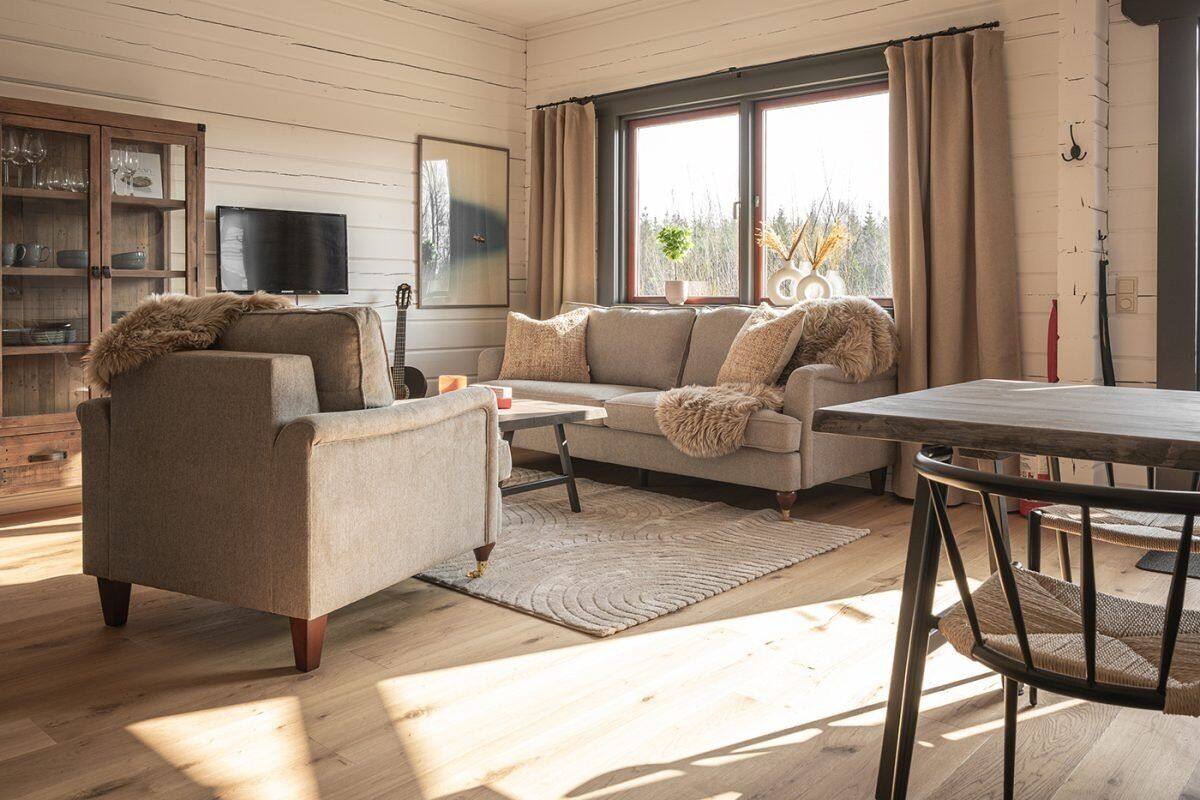Ein gemütliches Wohnzimmer mit einer Couch, einem Sessel und einem Couchtisch, einem Fernseher und einem Fenster mit Vorhängen.