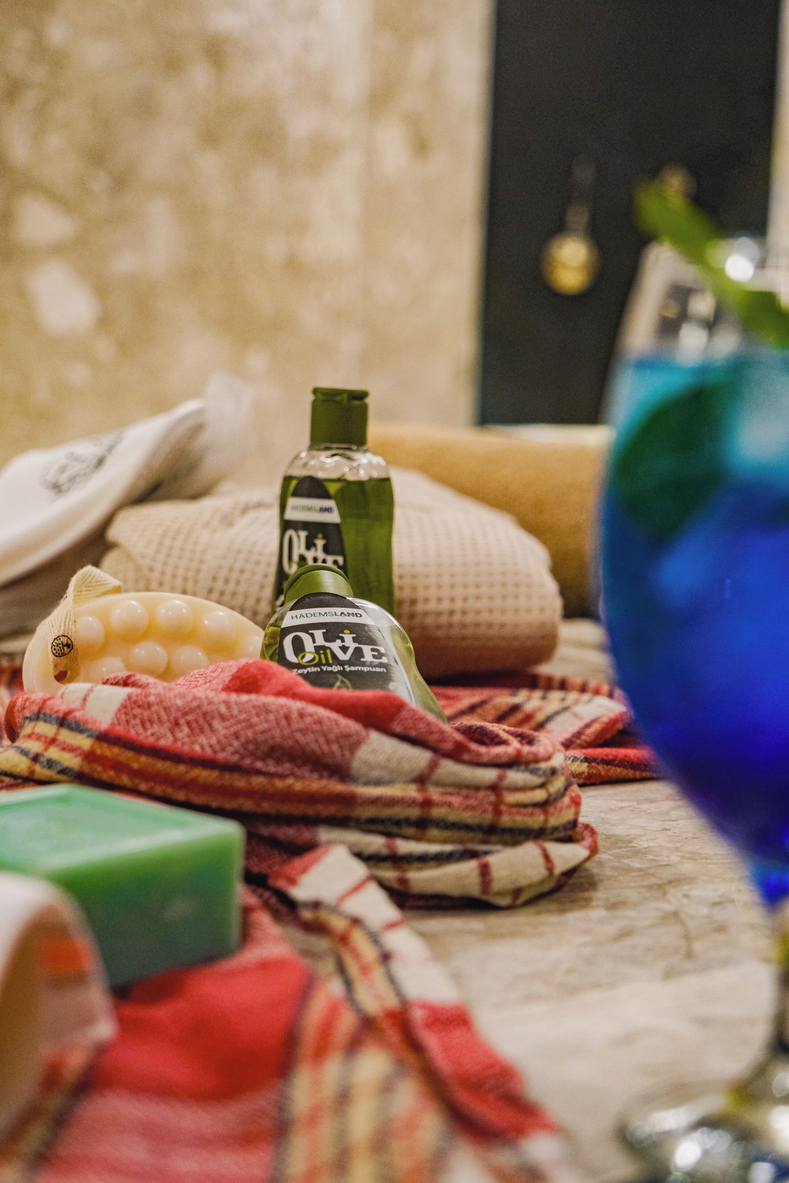 Una botella de aceite de oliva, una pastilla de jabón verde, un vaso azul y un paño escocés sobre una superficie texturizada.