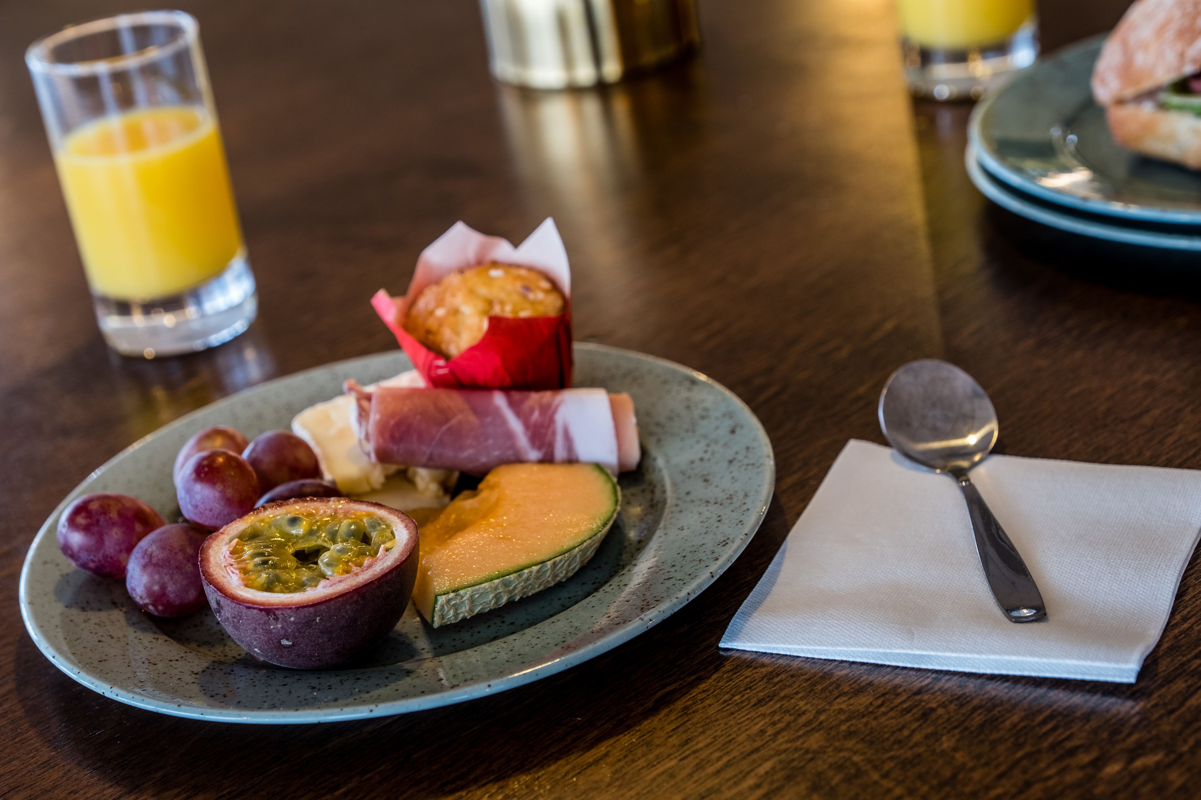 A plate with various fruits and a slice of melon, accompanied by a spoon and a napkin on a wooden table.