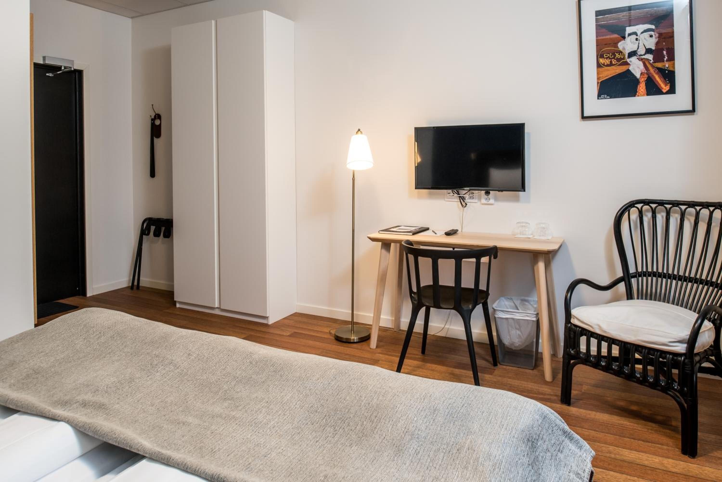 Une chambre d’hôtel comprenant un lit, un bureau avec une chaise, une télévision, un tableau encadré et une poubelle.