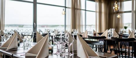 Un restaurant avec des tables dressées, de grandes fenêtres et des luminaires suspendus.