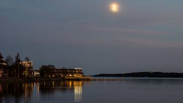 A waterfront hotel with a reflection in the calm water, a visible moon in the sky, and a distant shoreline.