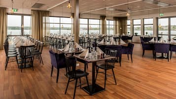 Un comedor con mesas dispuestas para el servicio, grandes ventanales con vistas a una masa de agua y lámparas colgantes.