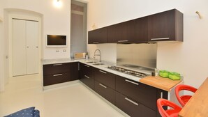Une cuisine moderne avec des meubles de couleur sombre, une cuisinière en acier inoxydable et un four encastré. Une petite fenêtre avec des stores et un téléviseur à écran plat fixé au mur complètent l’ensemble.