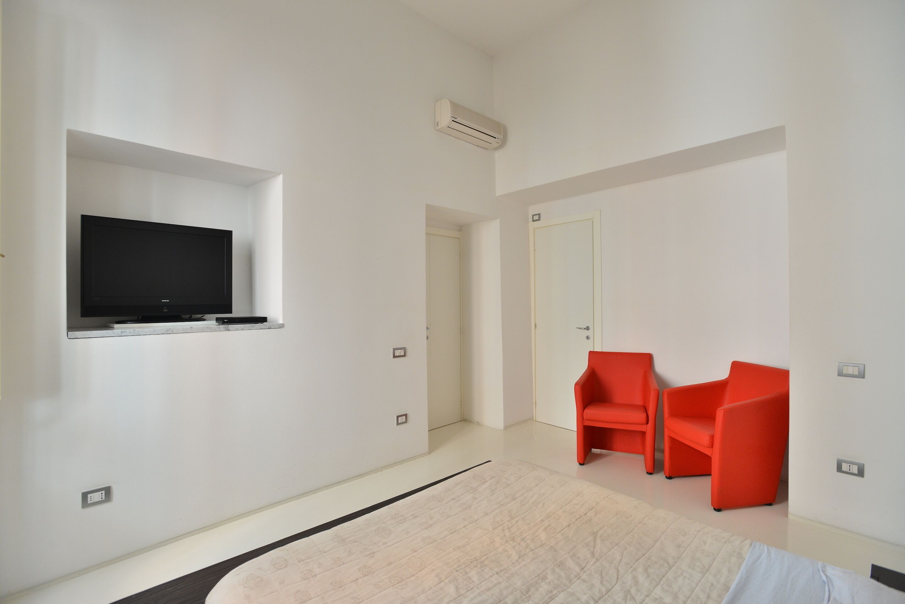 A modern hotel room with a flat-screen TV mounted on the wall, a door leading to another room, and two red chairs.