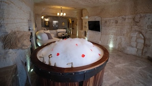 A stone-walled room with a large bathtub filled with water and rose petals, a sofa, and a television mounted on the wall. - Urgup Evi Cave Hotel (Ürgüp)