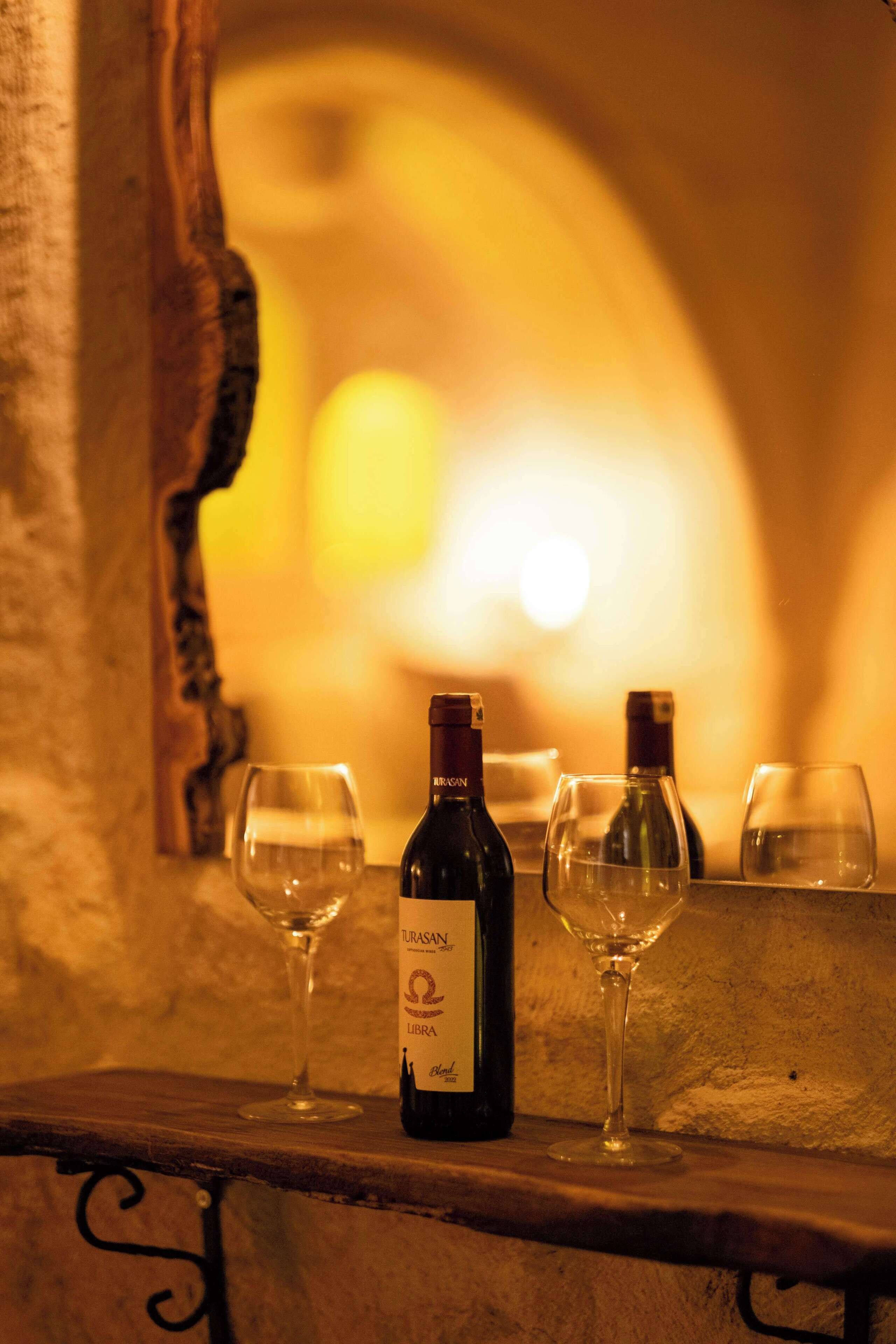 A bottle of wine and two glasses on a wooden shelf in a warmly lit room with a stone wall.