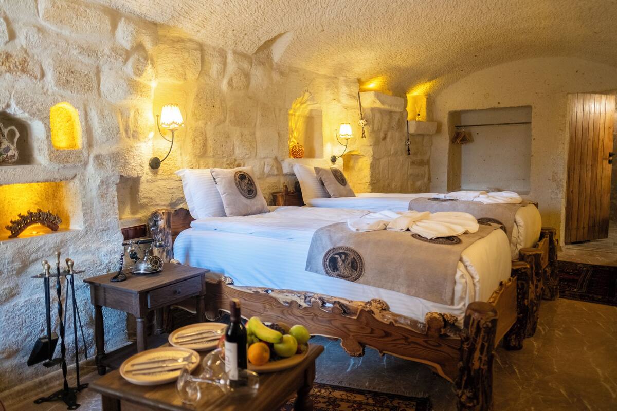 Un dormitorio rústico con paredes de piedra, dos camas, una mesa de madera con platos y una botella, y luces fijadas a la pared.