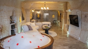 A spa room with a stone interior, a large bathtub with rose petals, a sofa, and a television. - Urgup Evi Cave Hotel (Ürgüp)