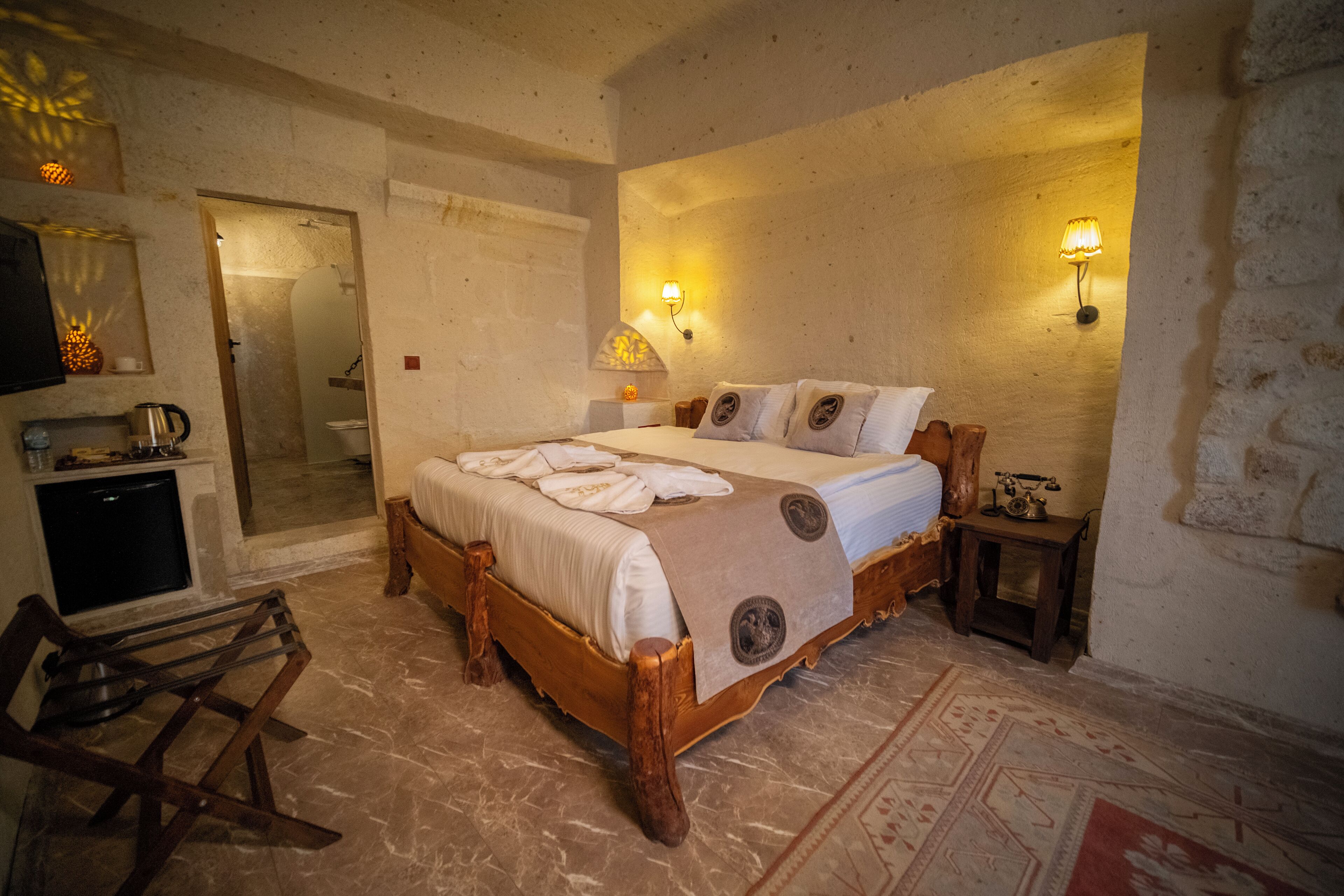 A bedroom with a wooden bed, two wall-mounted lamps, a small table with a lamp, a mirror, and a wooden chair.