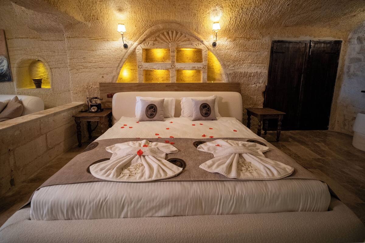 Un dormitorio acogedor con pared de piedra, cabecera de madera y cama con sábanas blancas y pétalos de rosa.
