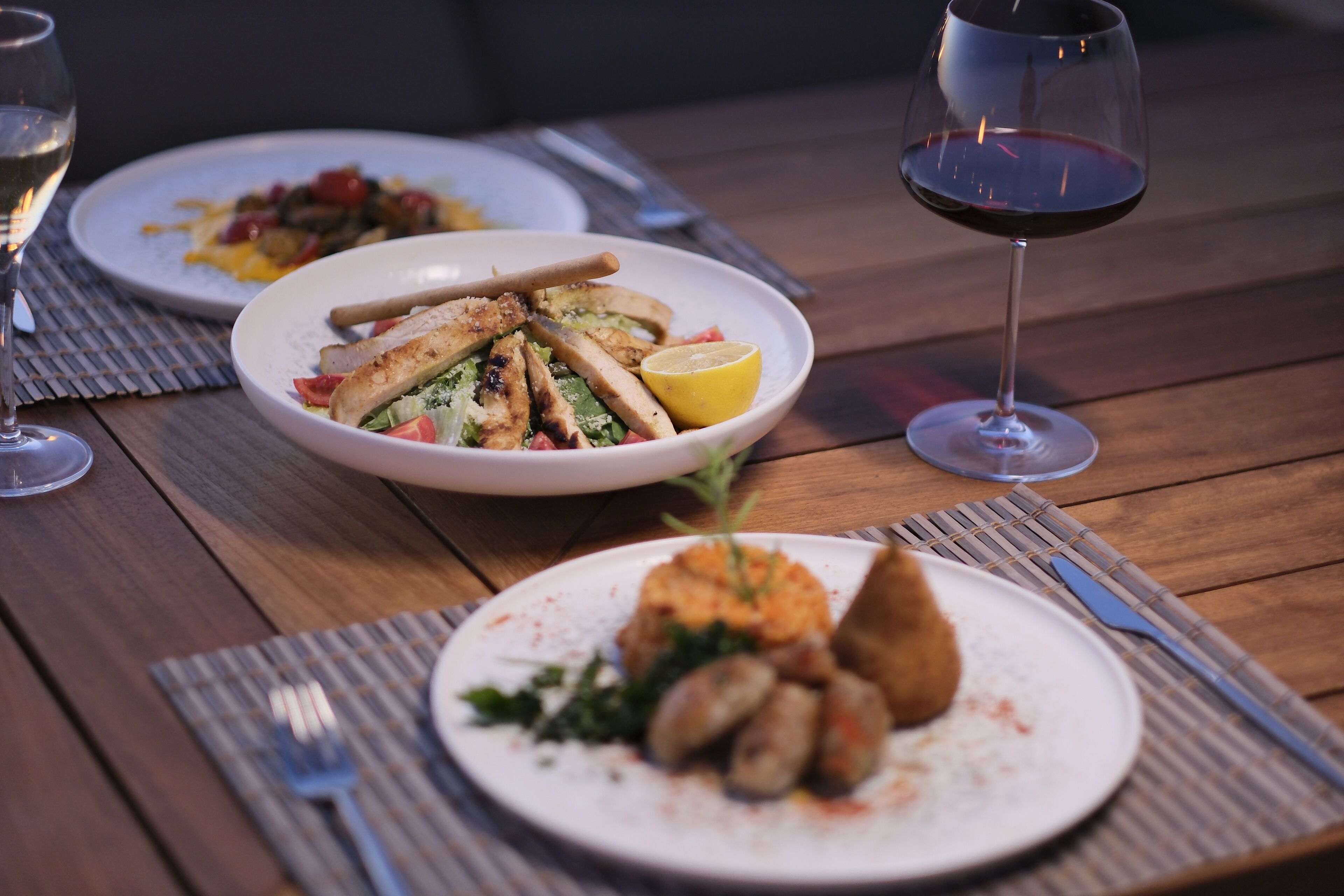 A wooden table with three plates of food and two glasses of wine.