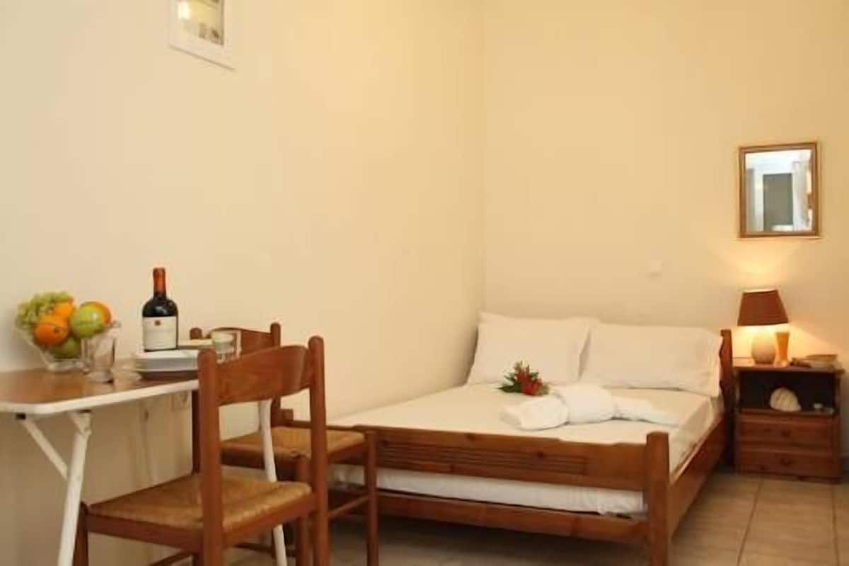 A small, cozy room with a wooden bed, a small table with a bottle and fruit, and a nightstand with a lamp.