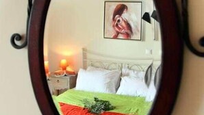 Un espejo con un marco decorativo refleja una cama con una colcha verde y roja, un almohadón blanco y un cuadro en la pared.