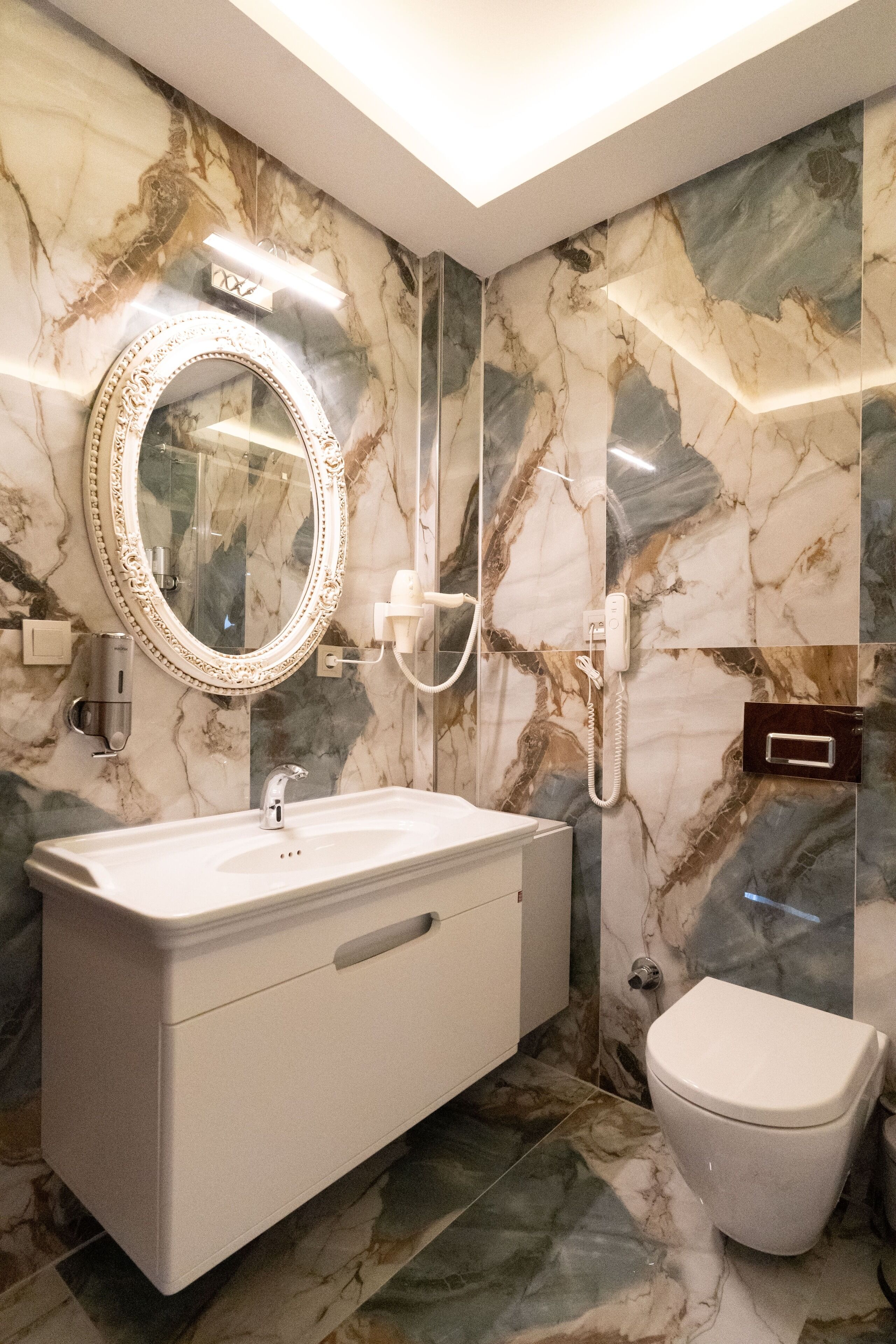 A bathroom with a marble wall, a sink, a toilet, and a mirror with a light above it.