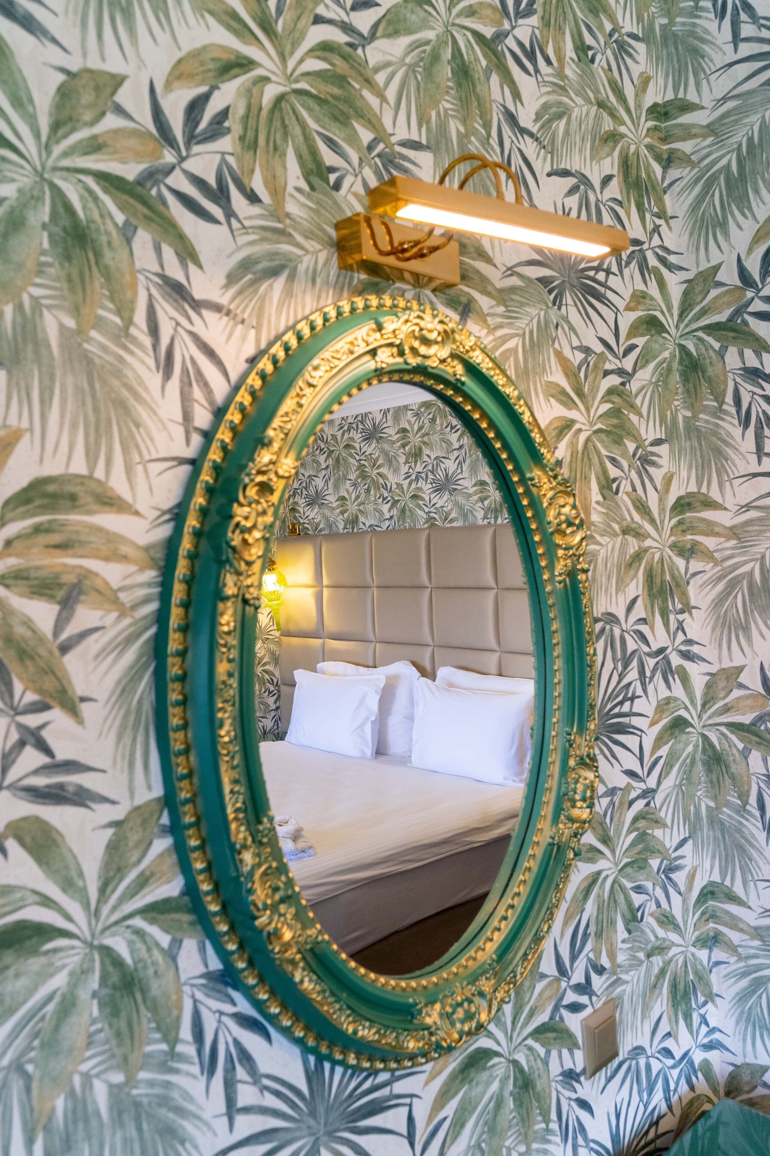 A mirror with an ornate gold frame, a bed with white linens, and a wall with a tropical leaf pattern.
