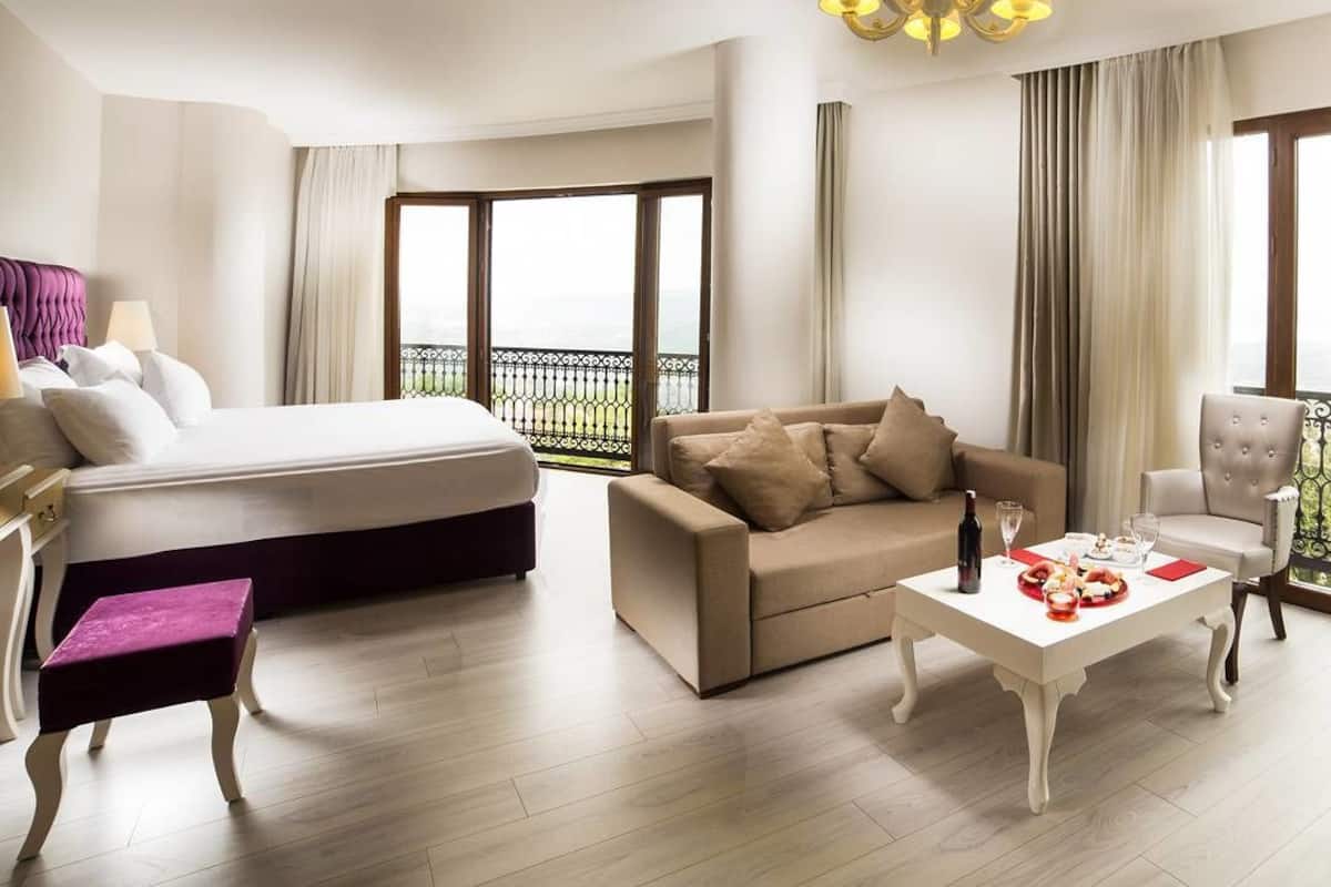 Habitación de hotel con cama, sofá, mesita de centro y una silla. Tiene un balcón con vistas.