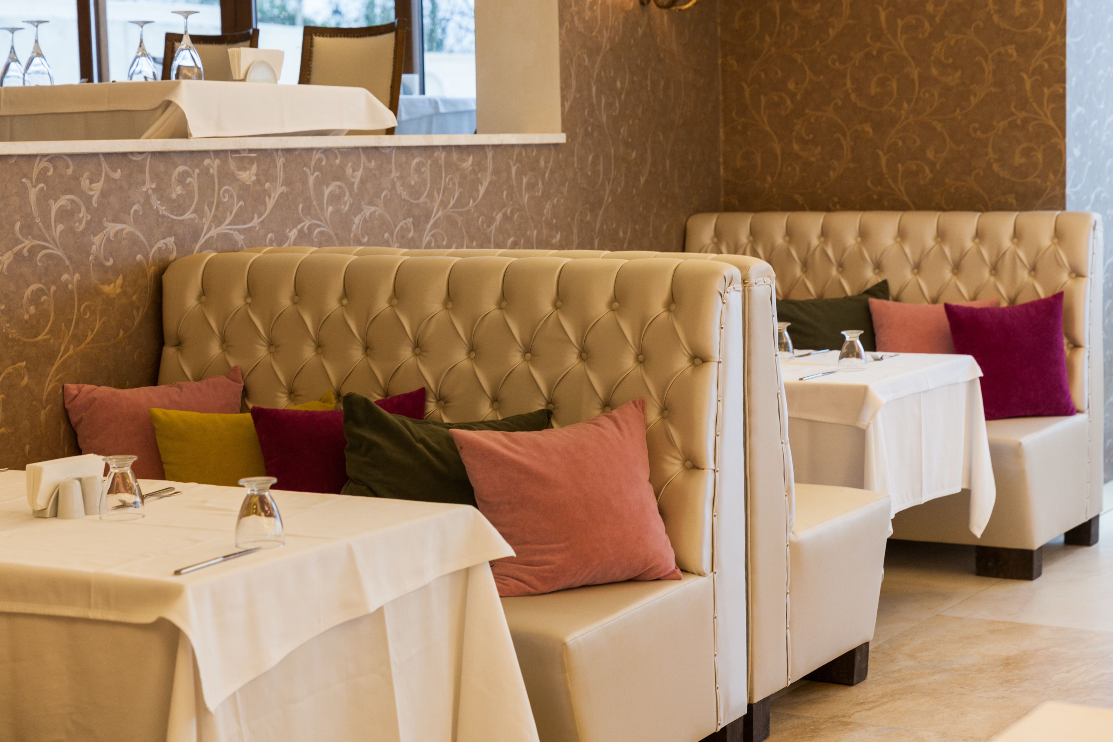 A restaurant with a tufted sofa and cushioned chairs, white tablecloths, and a patterned wall.