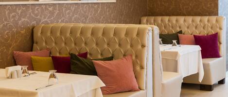 A restaurant with a tufted sofa and cushioned chairs, white tablecloths, and a patterned wall.