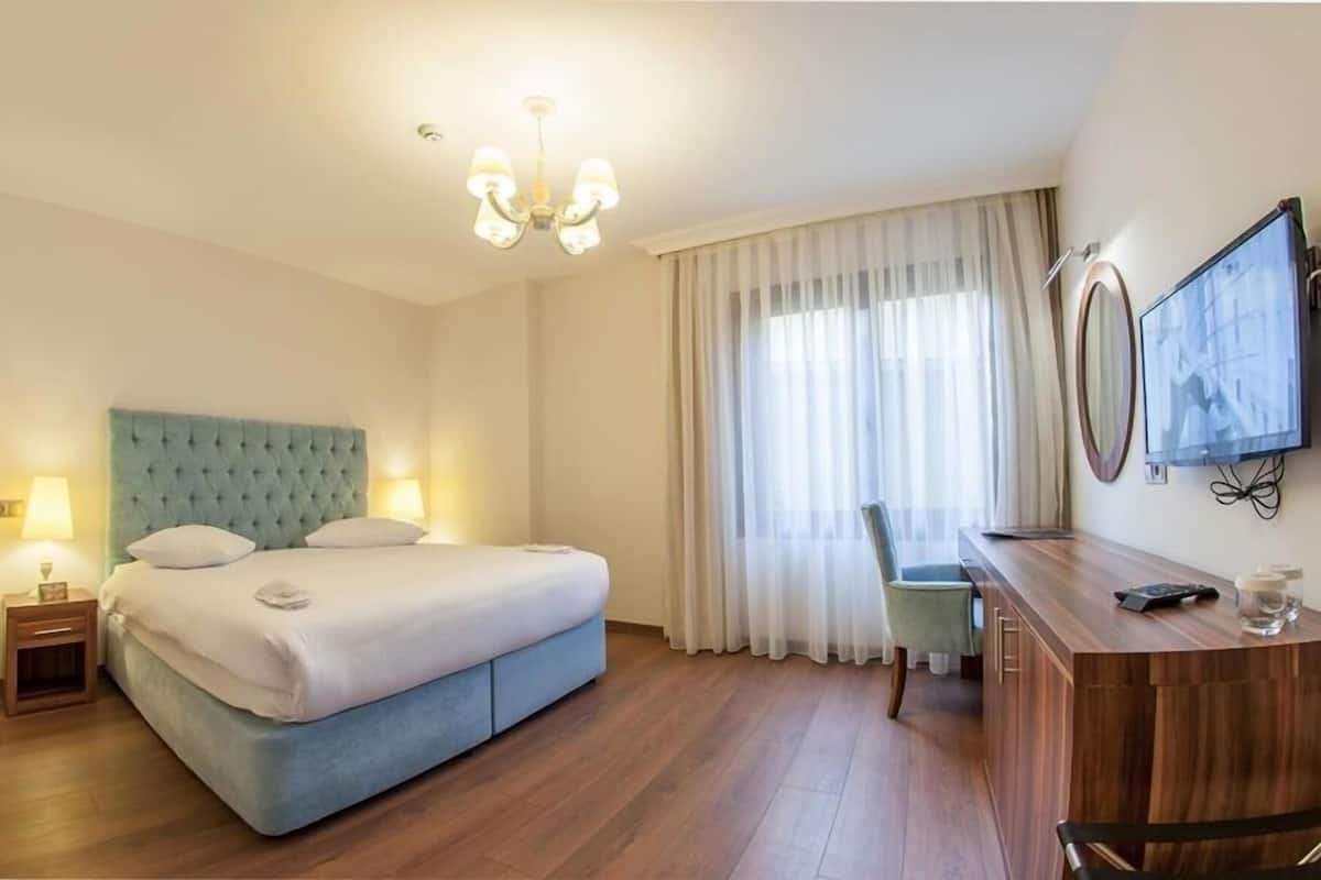 Habitación de hotel con cama, lámparas de noche, escritorio con silla, televisor y una ventana grande con cortinas.