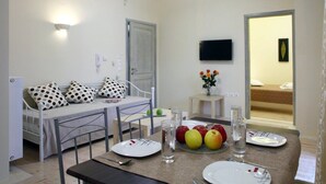 A dining area with a table set for two, a bed with a polka dot pillow, a small table with a vase of flowers, and a TV mounted on the wall. - Niriides (Lefkada)
