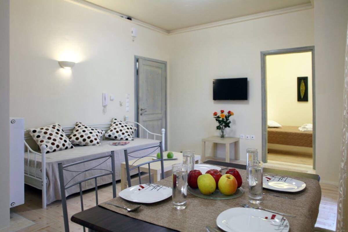 A dining area with a table set for two, a bed with a polka dot pillow, a small table with a vase of flowers, and a TV mounted on the wall.