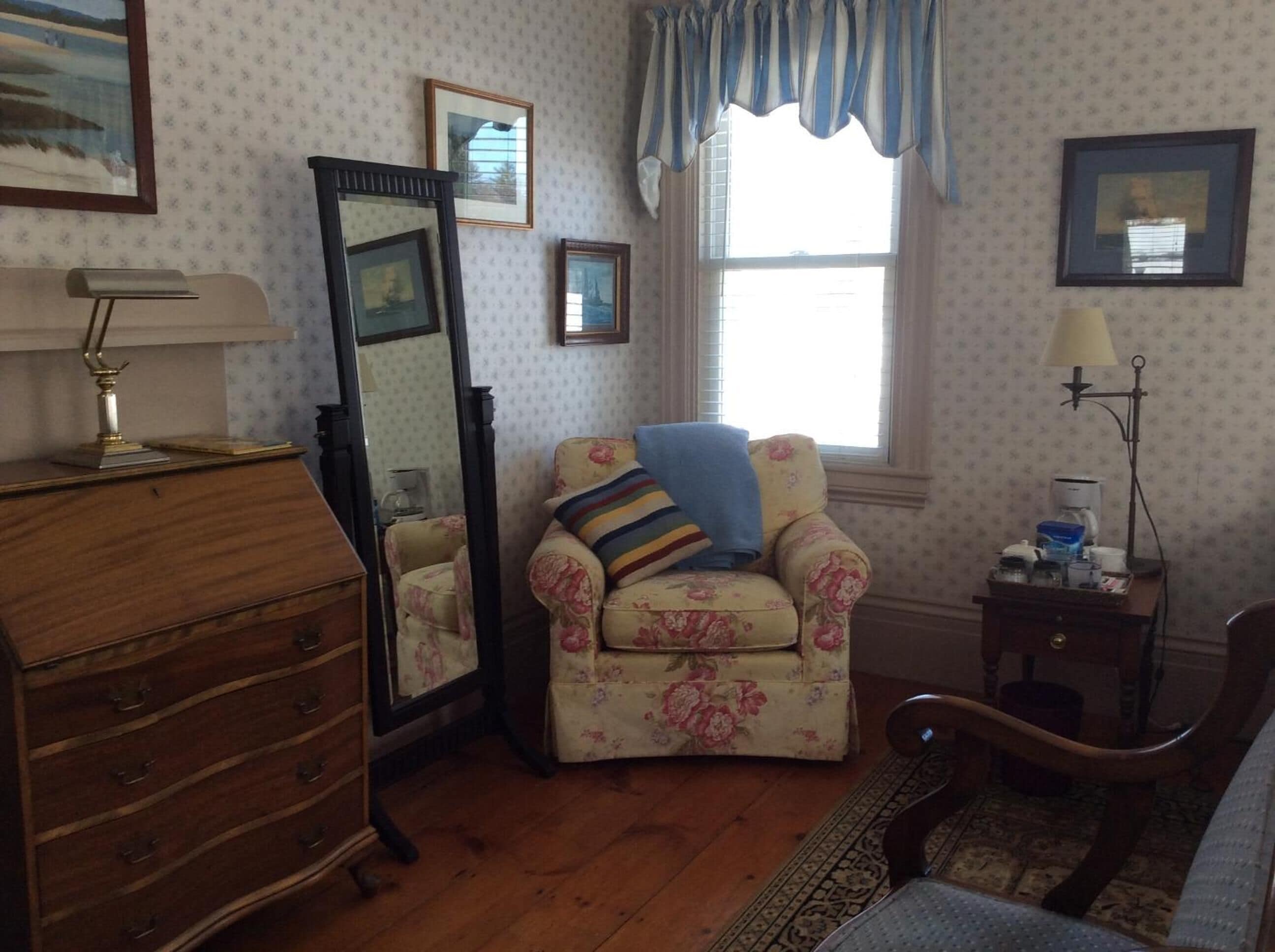 A room with a wooden dresser, a chair with a floral pattern, a mirror, and a window with blue curtains.