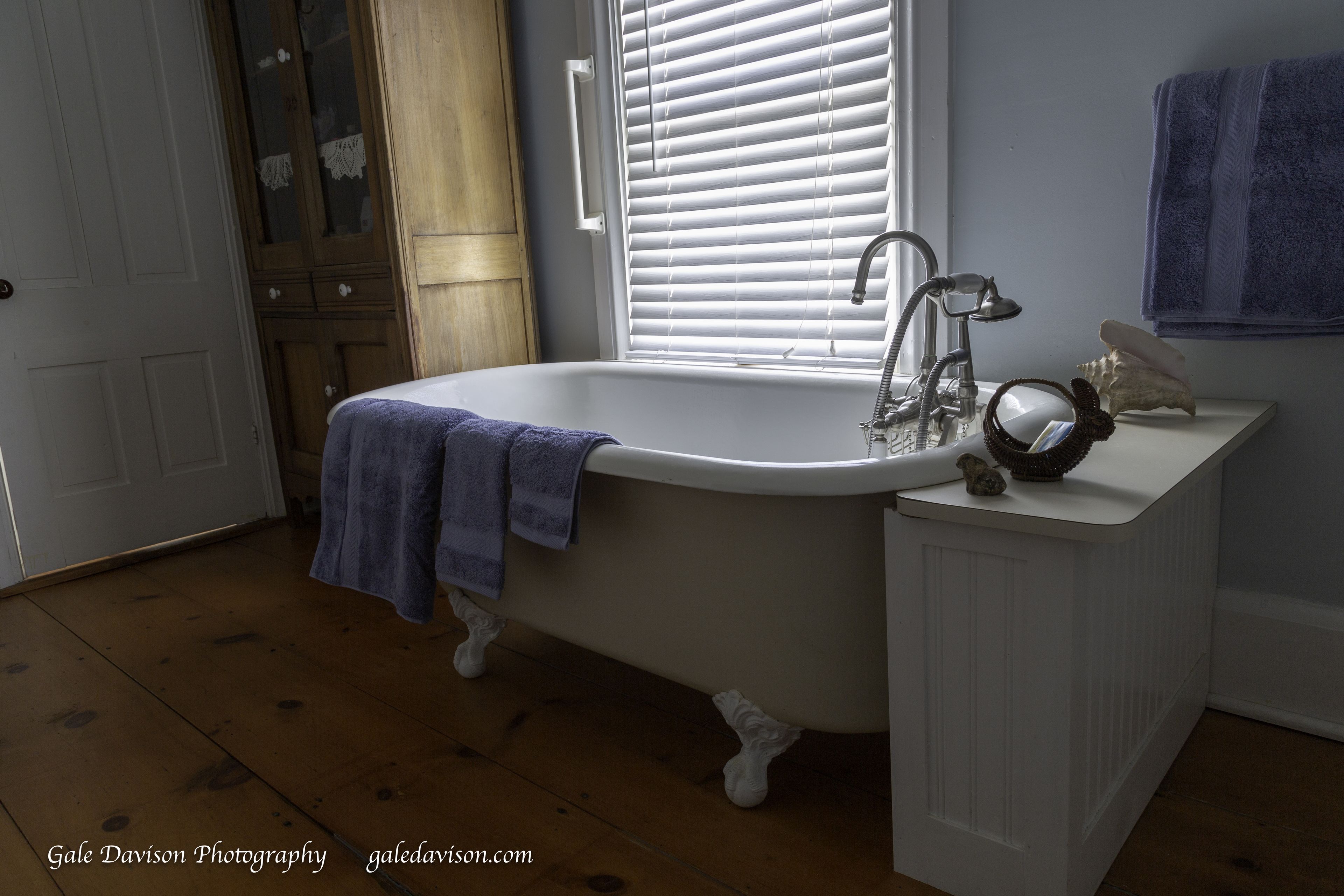 A bathroom with a white bathtub, a window with blinds, a sink with a mirror, and a towel rack.