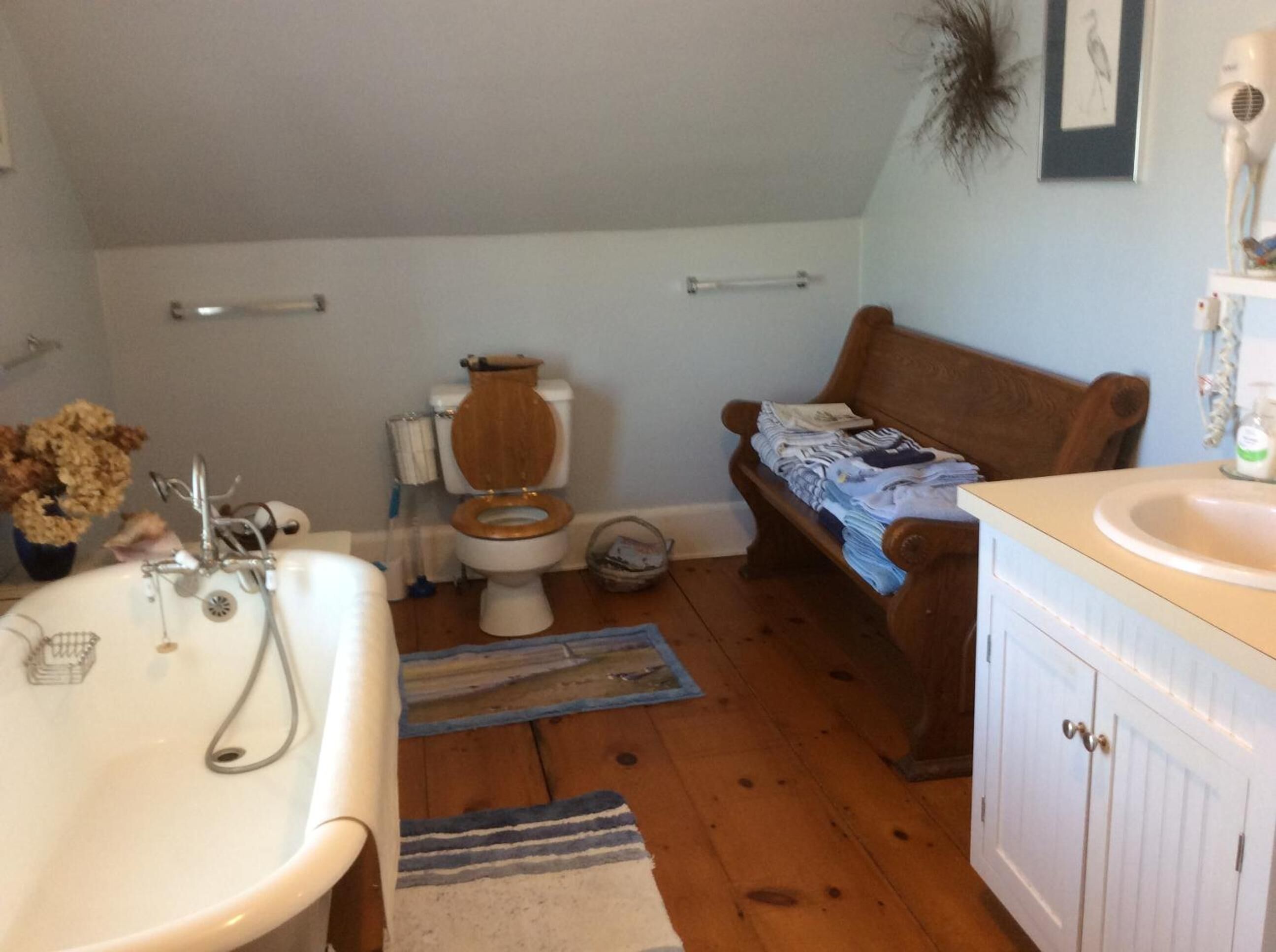 Une salle de bain avec des toilettes en bois, un lavabo blanc et un banc en bois sur lequel sont pliés des vêtements.