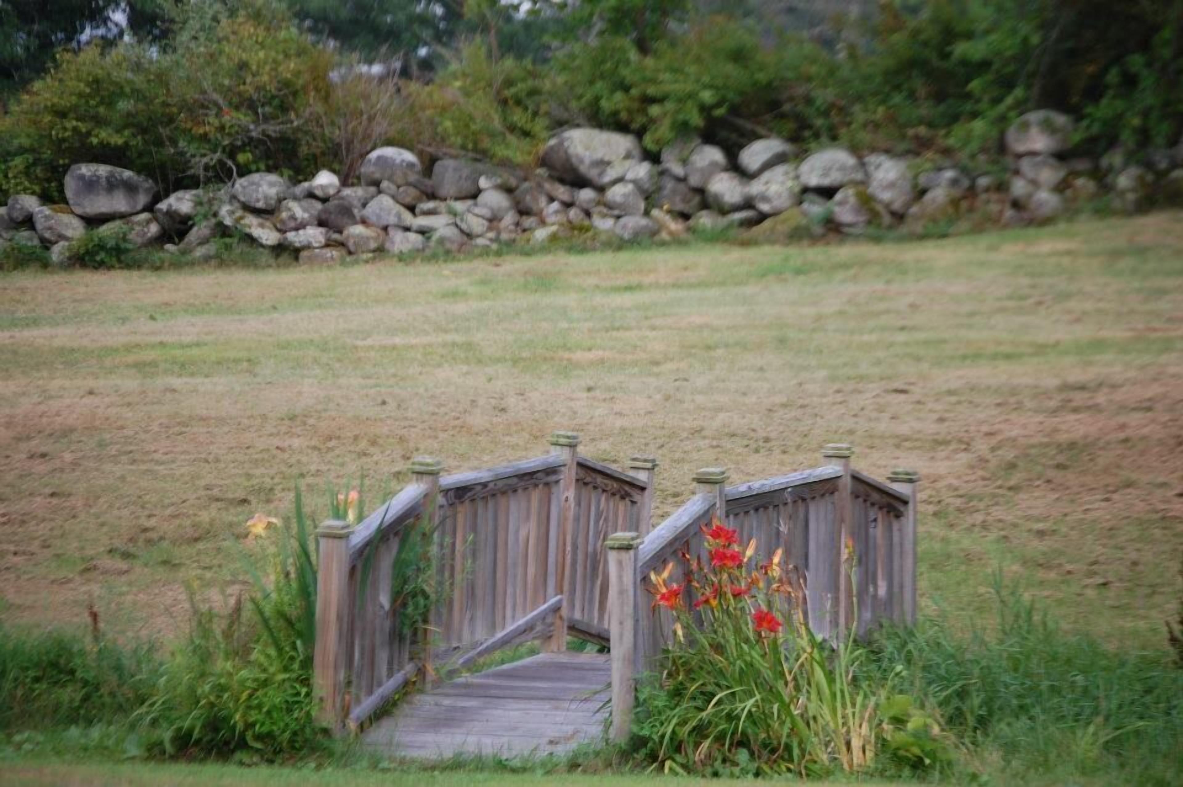 A wooden bridge in a grassy area with a stone wall in the background.