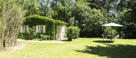 Un jardín exuberante con una casa pequeña, una mesa de comedor con sillas y un paraguas grande.