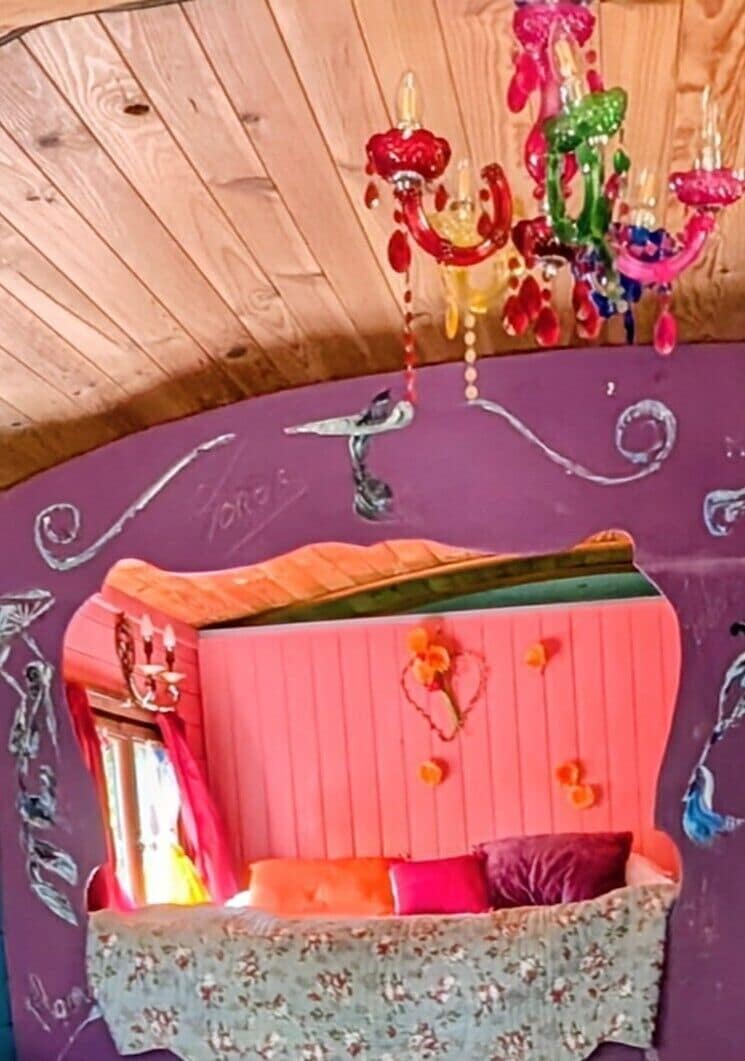 Ein Zimmer mit Holzboden, einer rosafarbenen Wand, einem Bett mit floralem Bettzeug und einem farbenfrohen Kronleuchter.