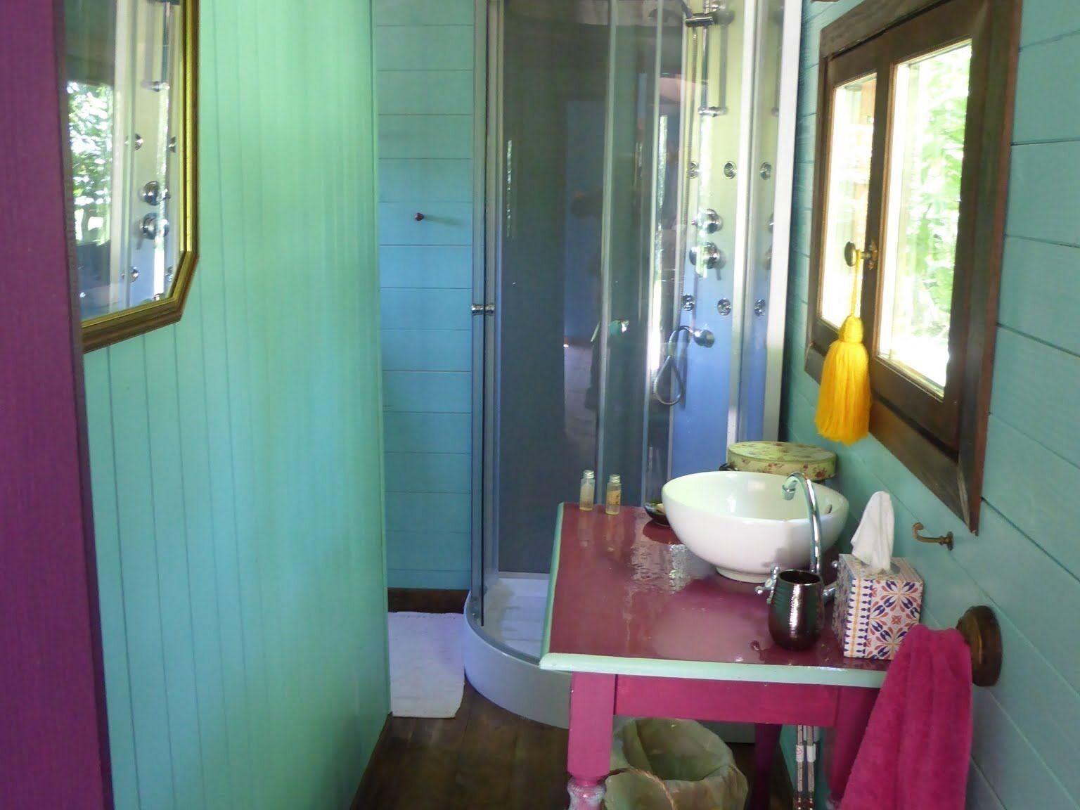 Une salle de bain avec une douche à parois de verre, un lavabo avec un miroir et une fenêtre ornée d’un rideau jaune.