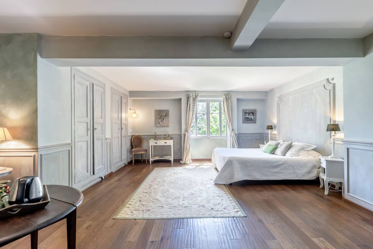 Quarto com cama grande, escrivaninha e cadeira. Há uma janela com cortinas, luminária e um quadro na parede.