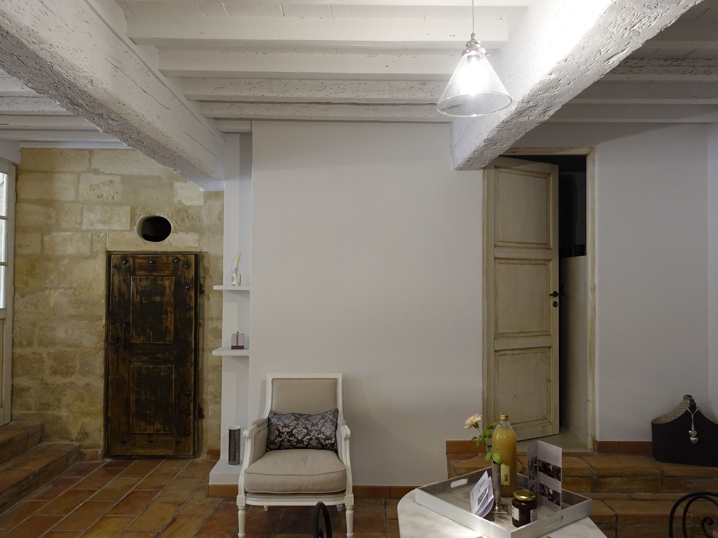 Une pièce avec un mur en pierre, une porte en bois, un fauteuil blanc, un luminaire suspendu et une petite table sur laquelle se trouvent une bouteille et un cadre photo.
