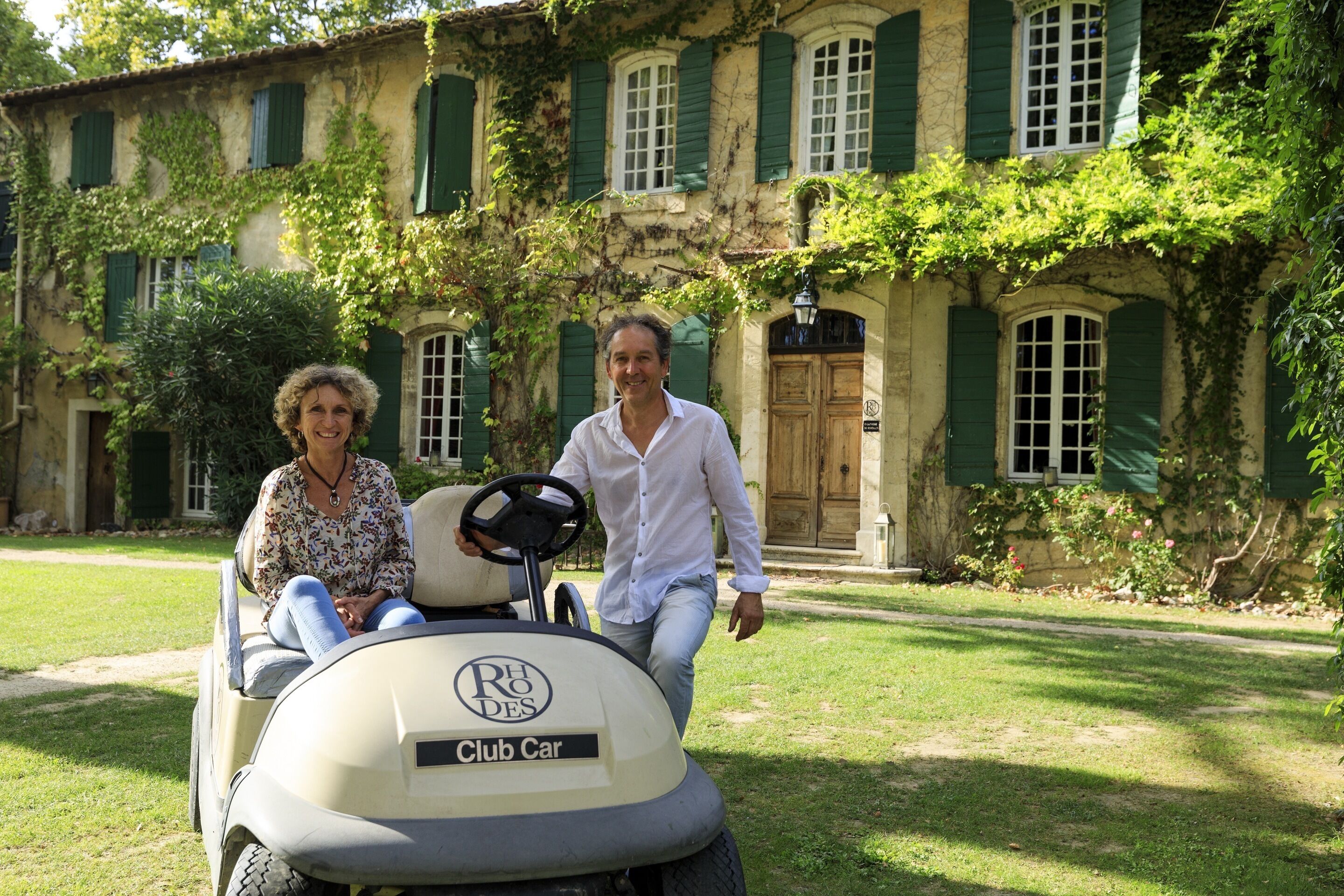 A golf cart with "RHO DES" and "Club Car" branding parked in front of a building with green shutters and ivy.