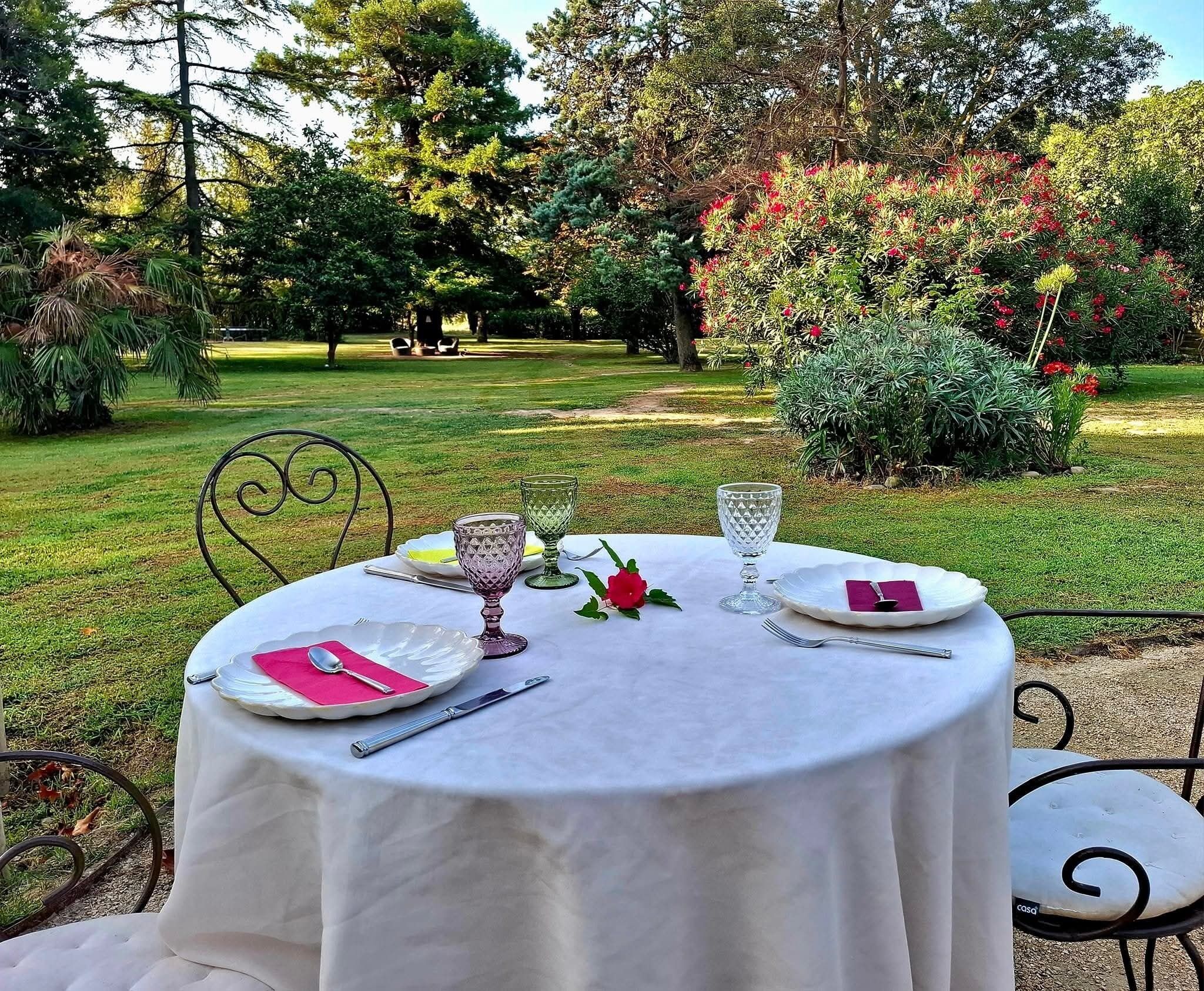 A round table set outdoors with white tablecloth, plates, cutlery, and a glass.