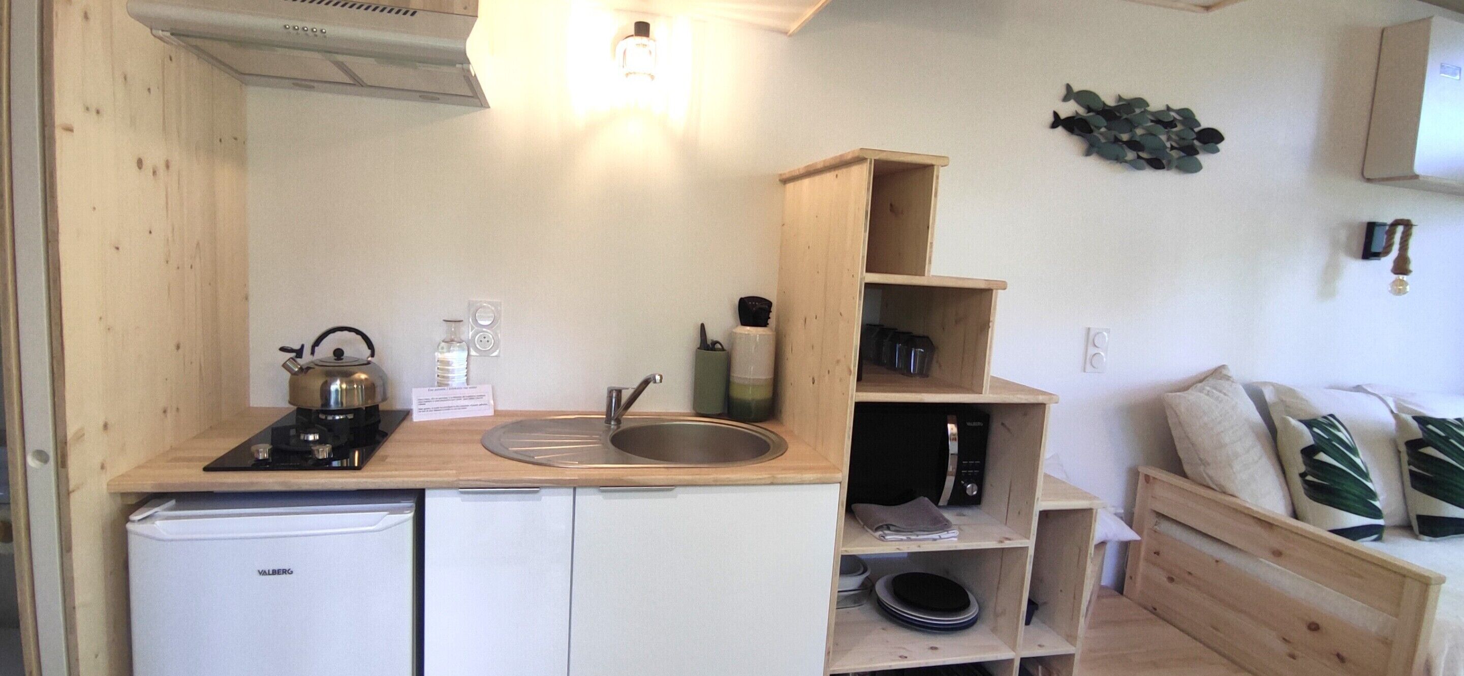 A compact kitchen with a sink, stove, and a small refrigerator. There are wooden shelves above the sink and a microwave on the right.