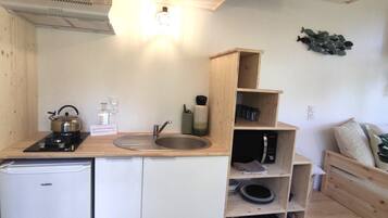 A compact kitchen with a sink, stove, and a small refrigerator. There are wooden shelves above the sink and a microwave on the right.