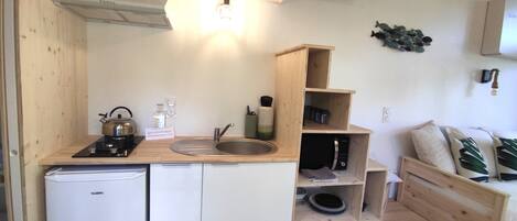 A compact kitchen with a sink, stove, and a small refrigerator. There are wooden shelves above the sink and a microwave on the right.