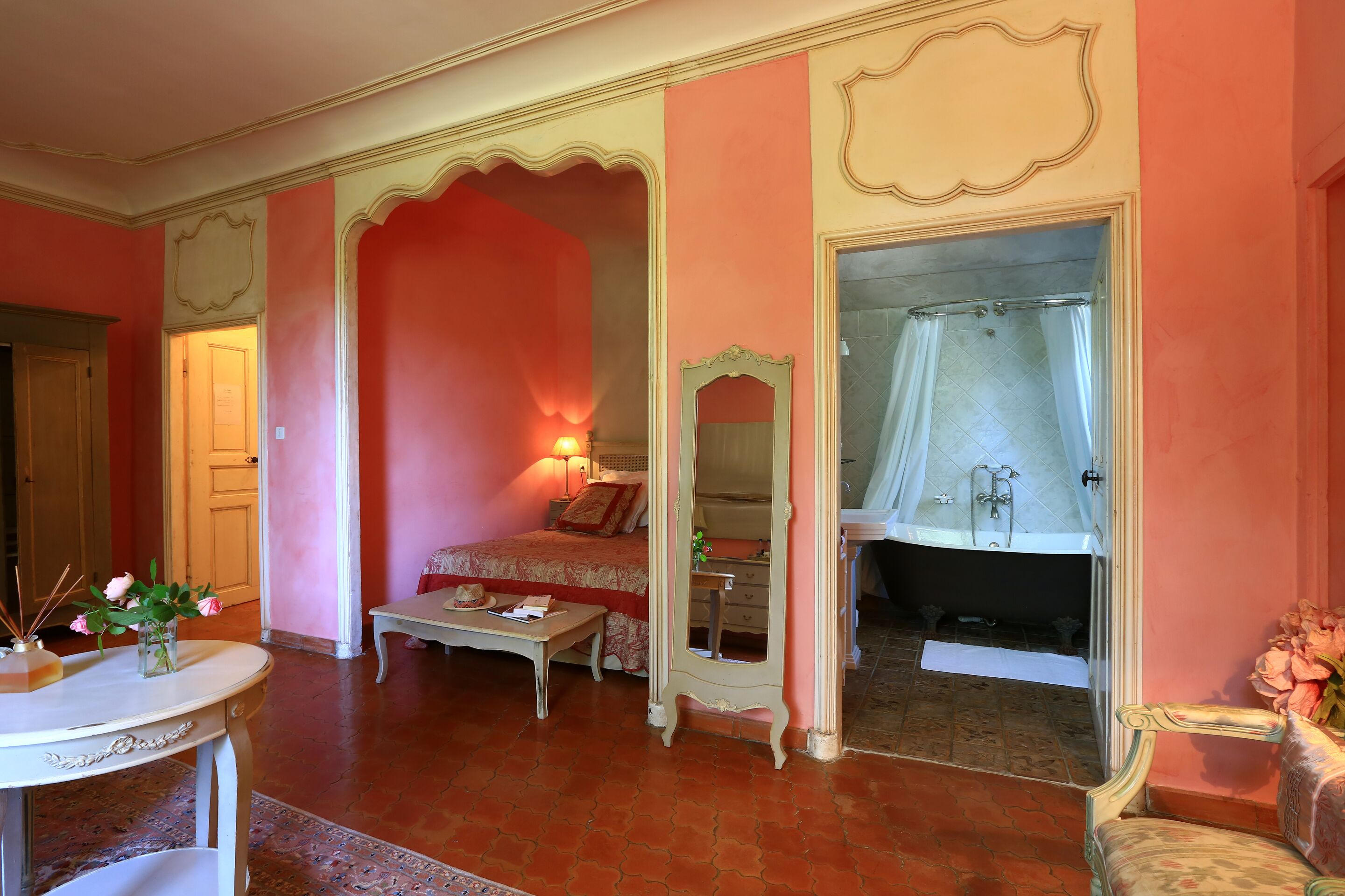 A room with a bed, a vanity with a mirror, a table with a vase of flowers, and a bathtub with a shower curtain.