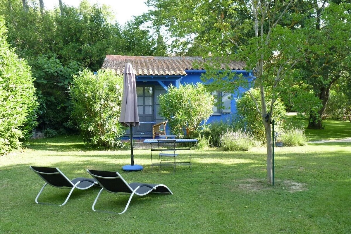 Uma casa azul com telhado de telhas, rodeada por um gramado bem cuidado e árvores. Há duas cadeiras de descanso e uma mesa pequena com guarda-sol.