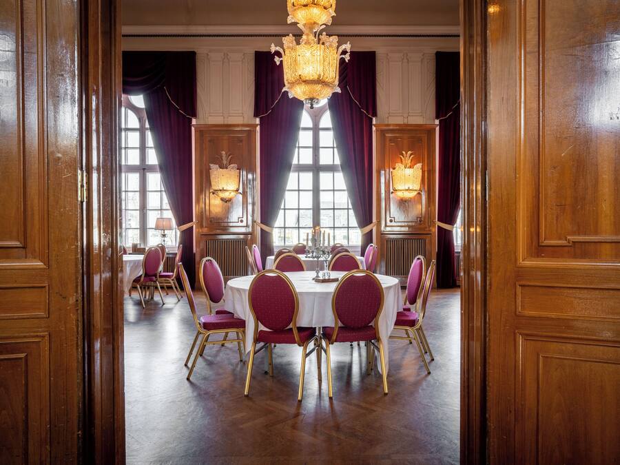 Une salle à manger élégante, avec des tables rondes, des chaises recouvertes de tissu rouge et des lustres.