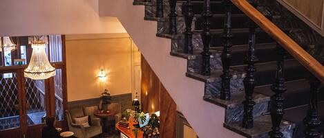 A grand staircase with black balusters and a wooden handrail, leading to an upper floor. A person is visible on the lower level, and a chandelier hangs from the ceiling.